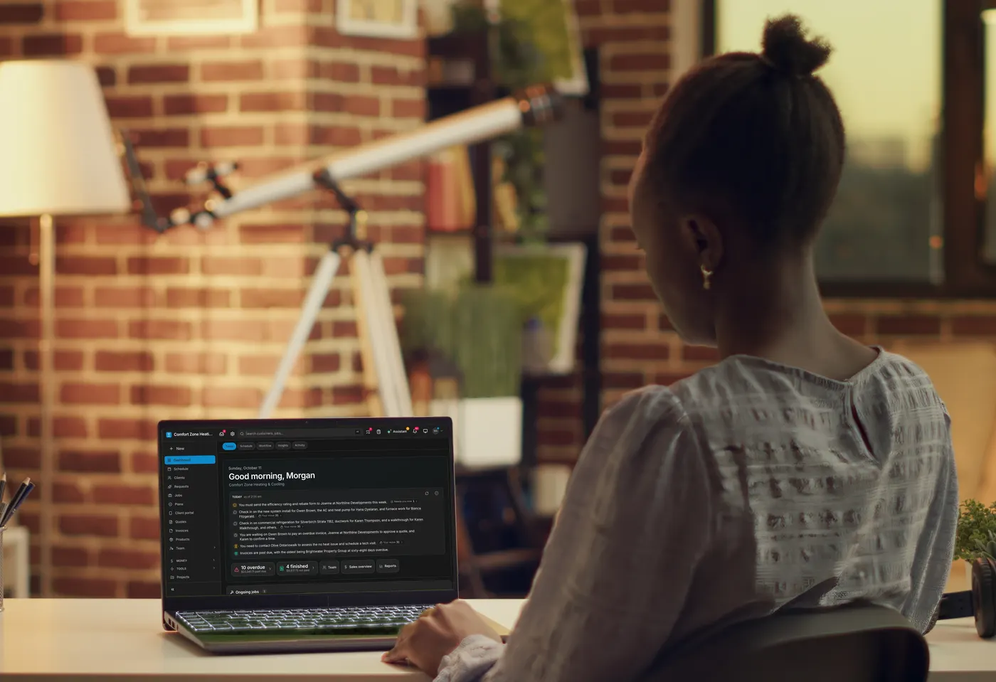 At dusk in a warm room, a woman works at a laptop on her desk: a sample heating and cooling company’s Daybook dashboard on its screen.